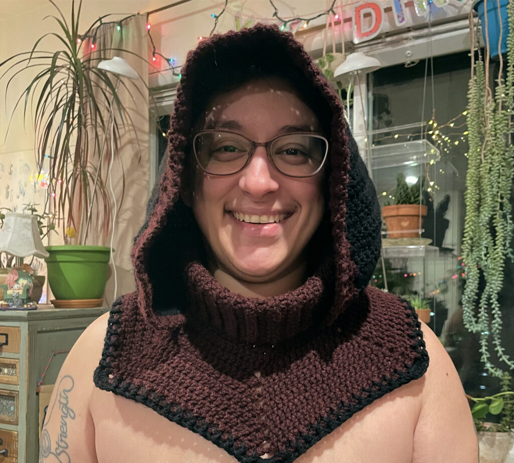 Danielle smiling at the camera wearing a dark red and black crochet hood with a collar around the neck that extends to the shoulders and chest. 